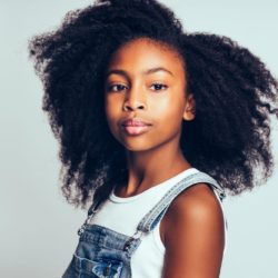 Confident young African girl with long curly hair and wearing dungarees standing by herself against a gray background